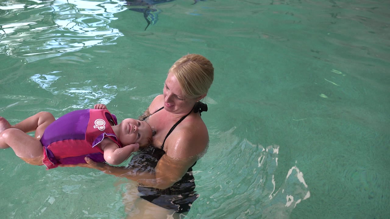 Woman holds a baby in a flotation vest in green pool water.