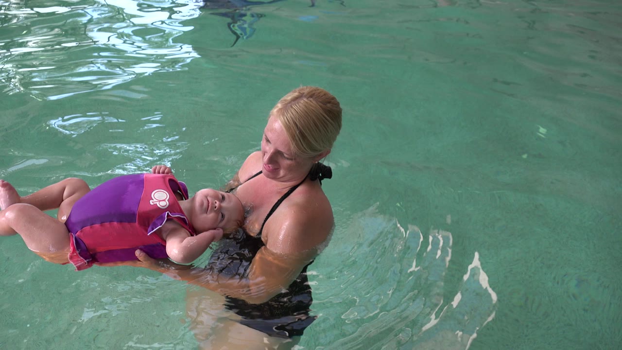 In Westminster, Colorado, the woman looks down toward the baby as the child floats on their back in a bright purple and pink flotation vest, surrounded by rippled green water.
