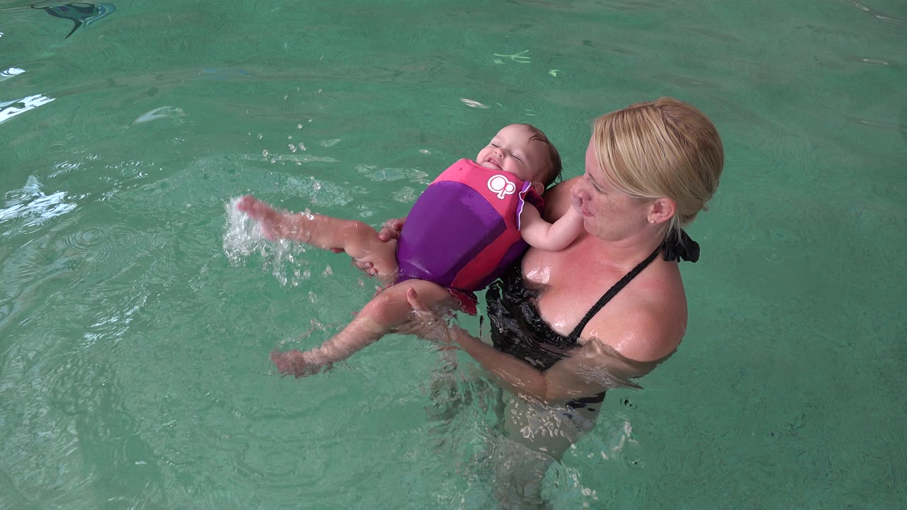 A woman in a black swimsuit supports a baby in a pink and purple flotation vest in a Westminster, Colorado pool as the baby's leg kicks water.