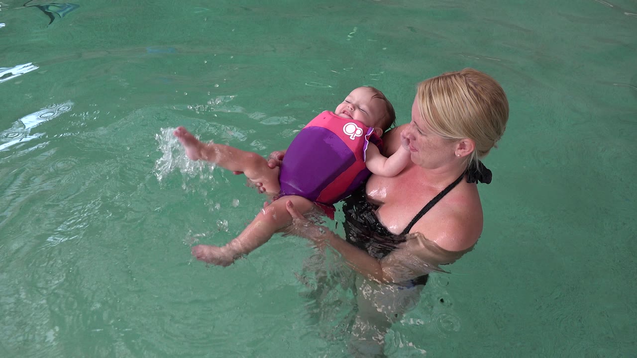 The baby leans back in the bright flotation vest while the woman holds them close in the Westminster, Colorado pool and a splash appears near the baby's foot.