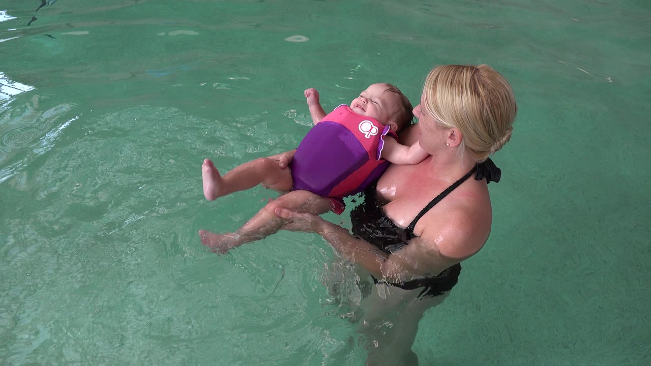 Woman lifts baby in flotation vest above green pool water.