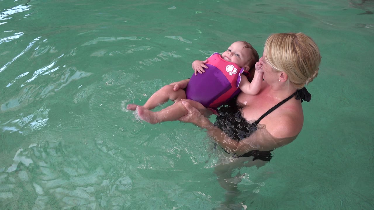 Mother holding a baby in a flotation vest in green pool water.