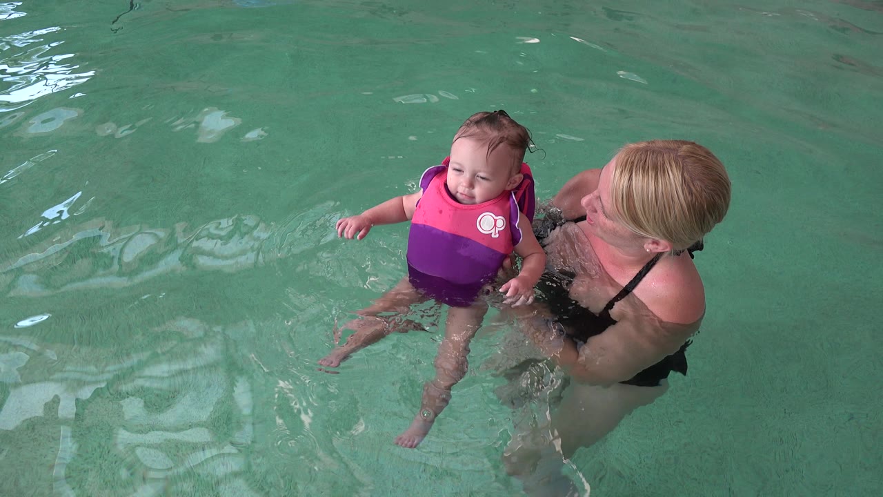 Mother supports a baby in a pink and purple flotation vest in pool water.