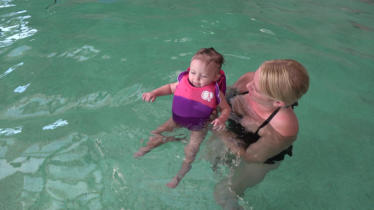 At 0:01 in Westminster, Colorado, the baby smiles while floating near the blonde woman, with rippled green water filling the frame.