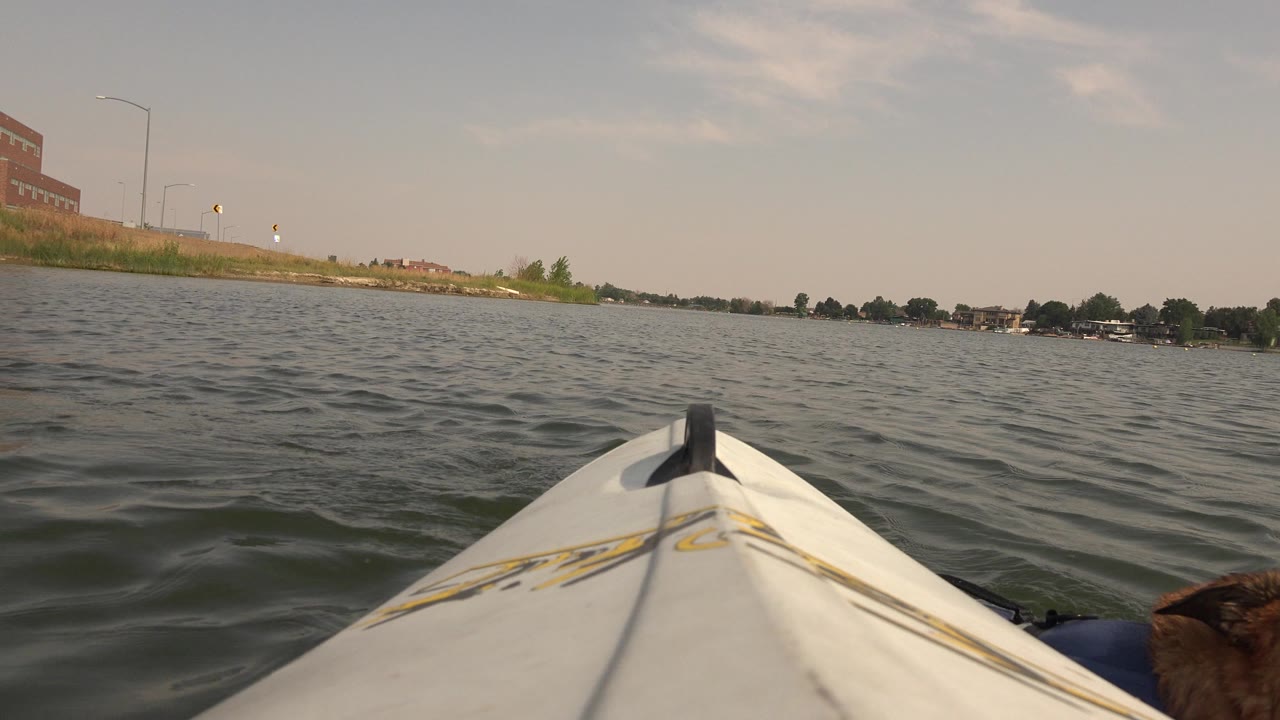 At 0:00, the white kayak bow points across Hidden Lake in Colorado toward a distant tree-lined shore with lakeside buildings and no visible people.