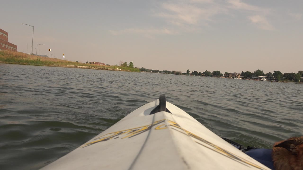 At 0:01, the kayak bow remains centered over small waves on Hidden Lake, with low buildings and trees along the far shore and no visible people.
