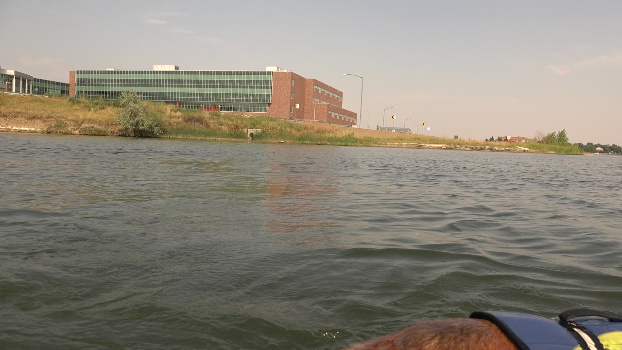 At Hidden Lake Colorado, the opening frame shows rippled water from a low kayak viewpoint, with part of a yellow-brown dog and safety jacket at the bottom edge, a grassy bank, a large brick-and-glass building, and no visible people.