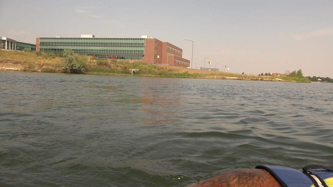 At 0:01 in Hidden Lake Colorado, the low water view keeps the large shoreline building and grassy bank in the background while the dog remains partly cropped near the bottom edge, with no visible people.