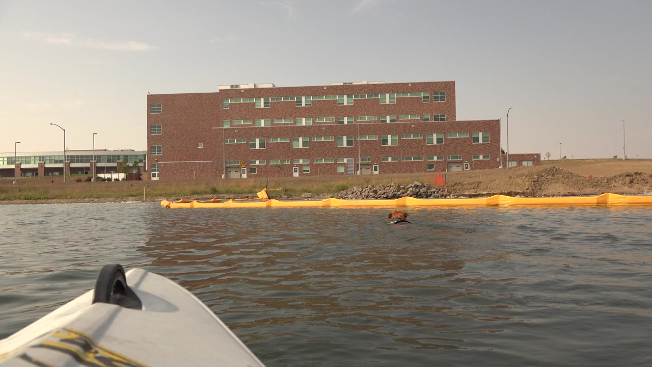 At Hidden Lake Colorado, a white boat bow points across rippled water toward a brown brick building and yellow floating barrier, with no visible people.