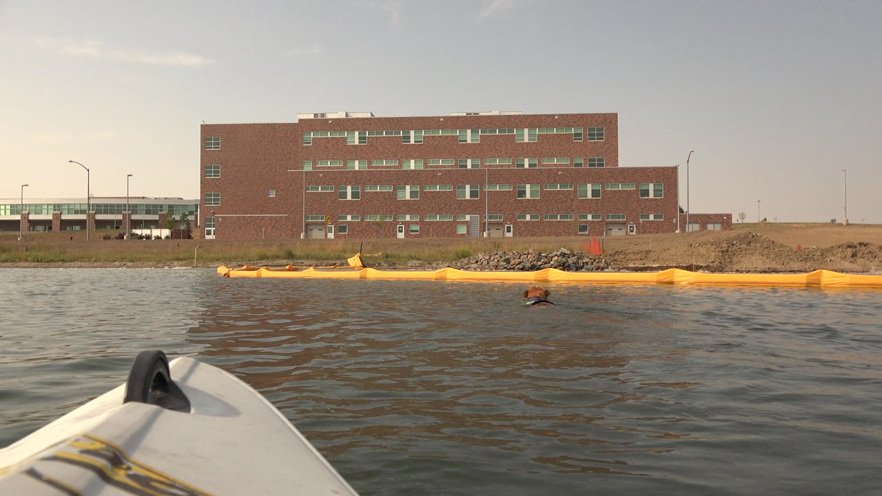 At Hidden Lake Colorado, the boat bow remains low in the frame as small waves cross the water in front of the yellow barrier and brick building, with no visible people.