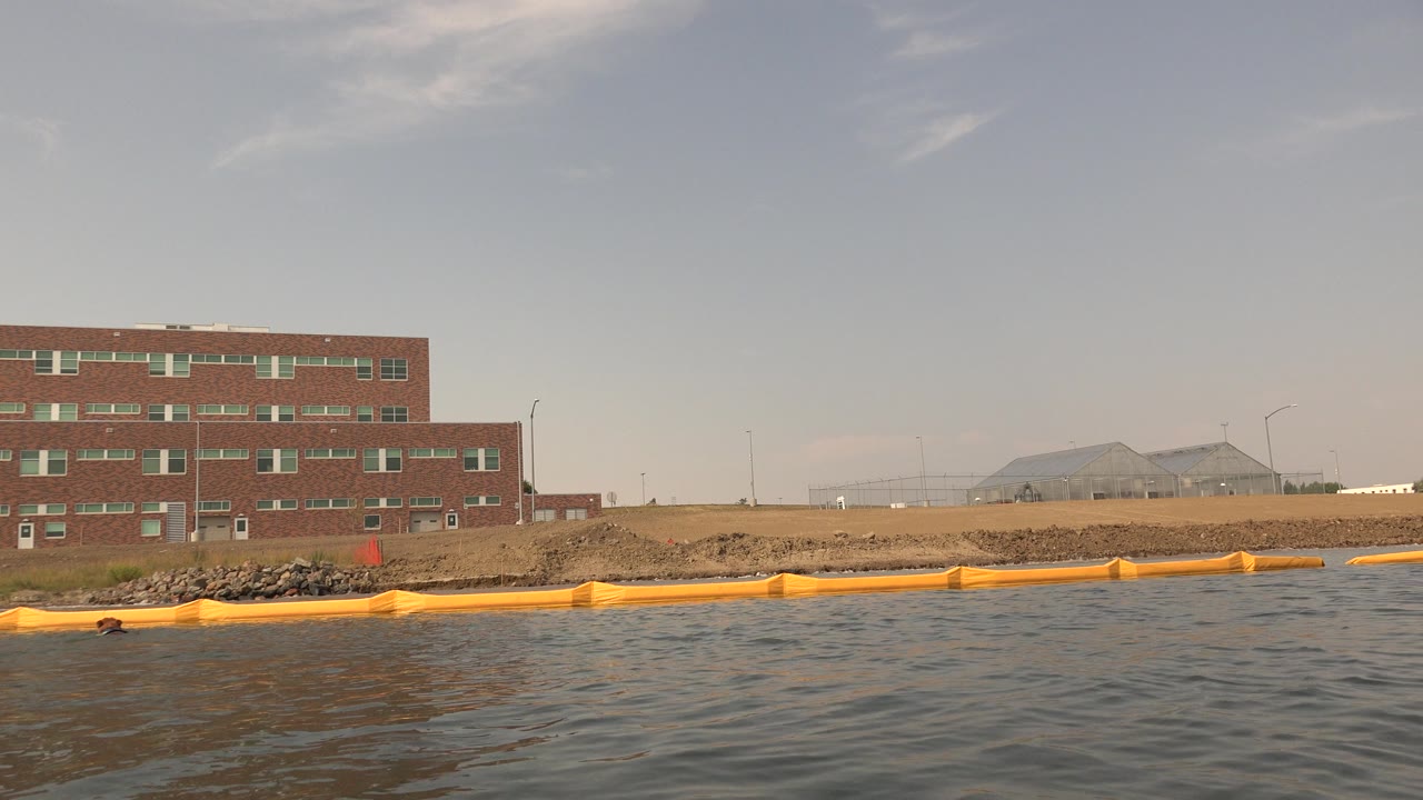 Hidden Lake Colorado frame with rippled water in front, a yellow floating barrier along the shore, a brown brick building at left, pale sky above, and no visible people.