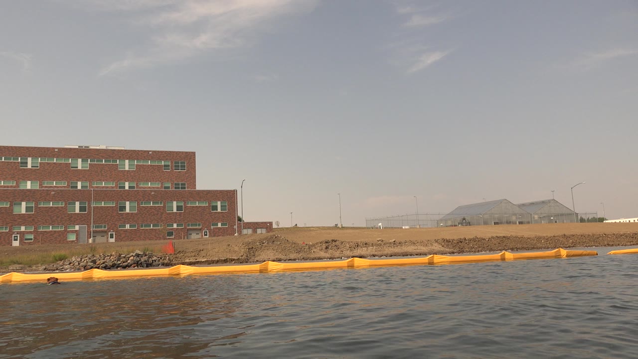 Hidden Lake Colorado view with the brick building more centered, the yellow barrier crossing the shoreline, textured lake water in the foreground, and no visible people.