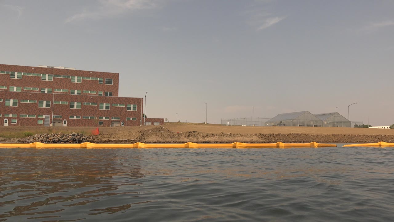 Low water-level view at Hidden Lake, Colorado, with rippled lake water in front, an orange floating boom along the shore, and a red brick building on the left.