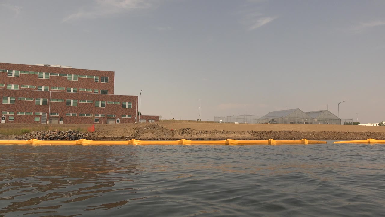 Hidden Lake, Colorado shoreline with dark rippled water, a continuous orange boom, a red brick building near center, and a pale arched structure farther back.