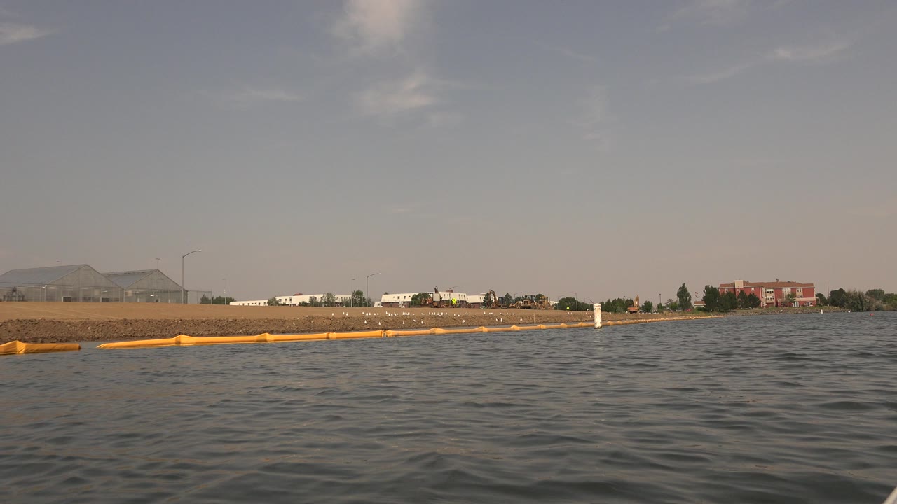 Hidden Lake Colorado shoreline seen over dark rippled water, with the yellow boom closer to the lower left and low buildings across the beach, no visible people.