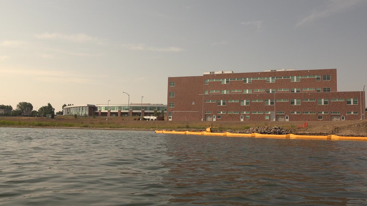 Wide view at Hidden Lake, Colorado with rippled water in the foreground, a yellow floating barrier near rocks, a brick building beyond the shore, and no visible people.