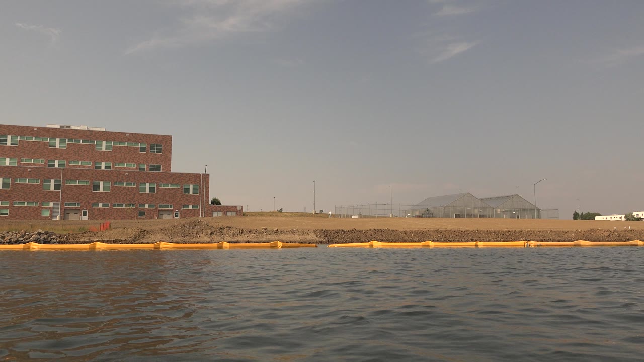 Opening water-level frame at Hidden Lake, Colorado showing rippled water, a yellow boom, rocky shoreline, a brick building, distant low hills, pale sky, and no visible people.