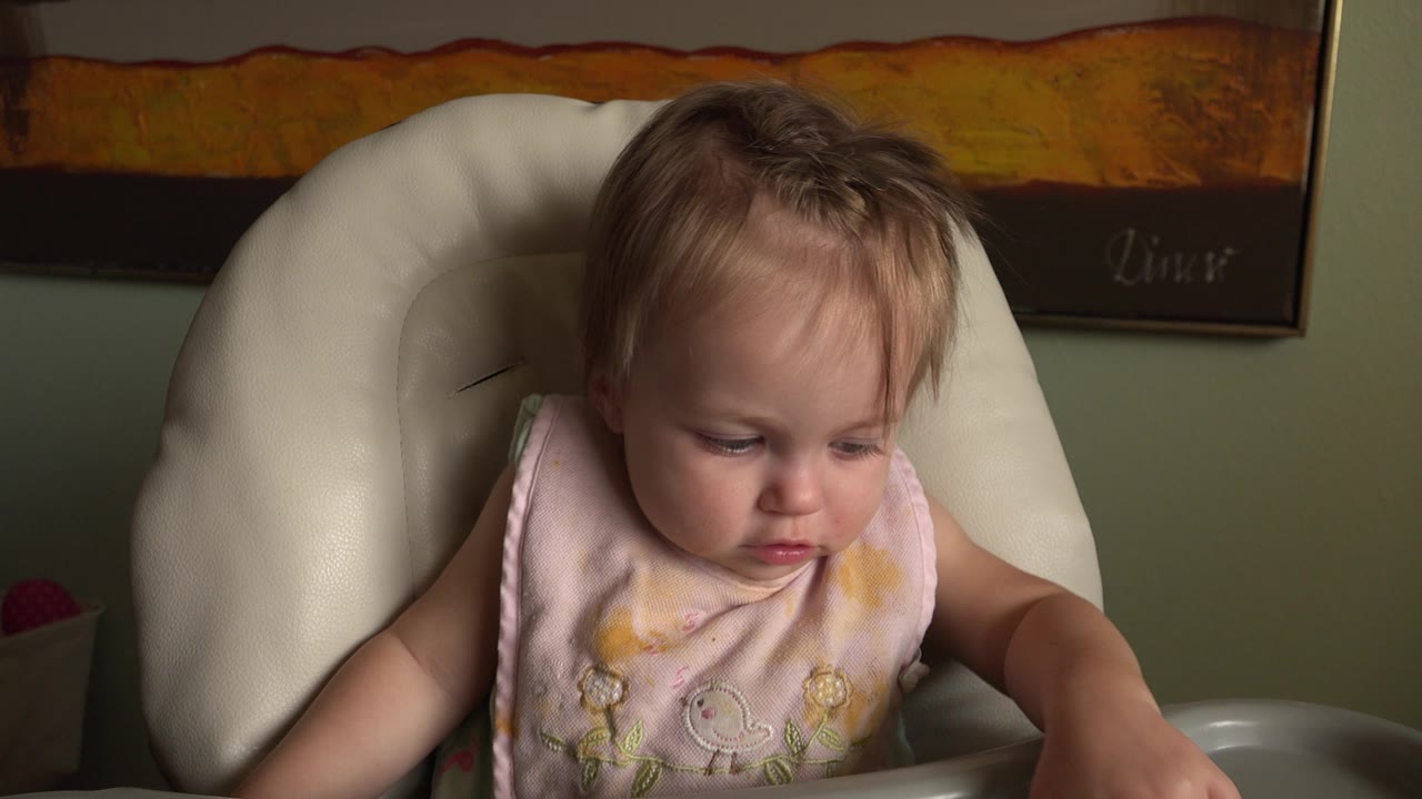 At Hidden Lake, Colorado, the baby remains seated in the high chair, head lowered and arm extended toward the tray in a dim indoor room.