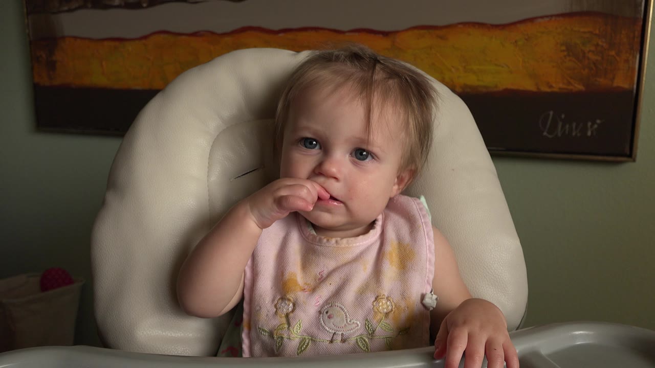 Infant girl in a high chair touching her mouth.