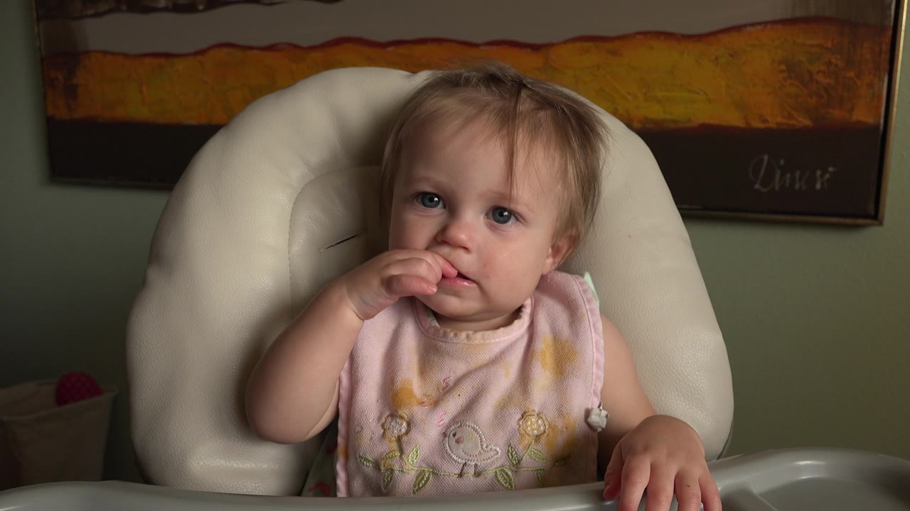 At 0:01, the infant remains centered in the white high chair, looking toward the camera while holding fingers at her mouth with the tray edge visible in front.