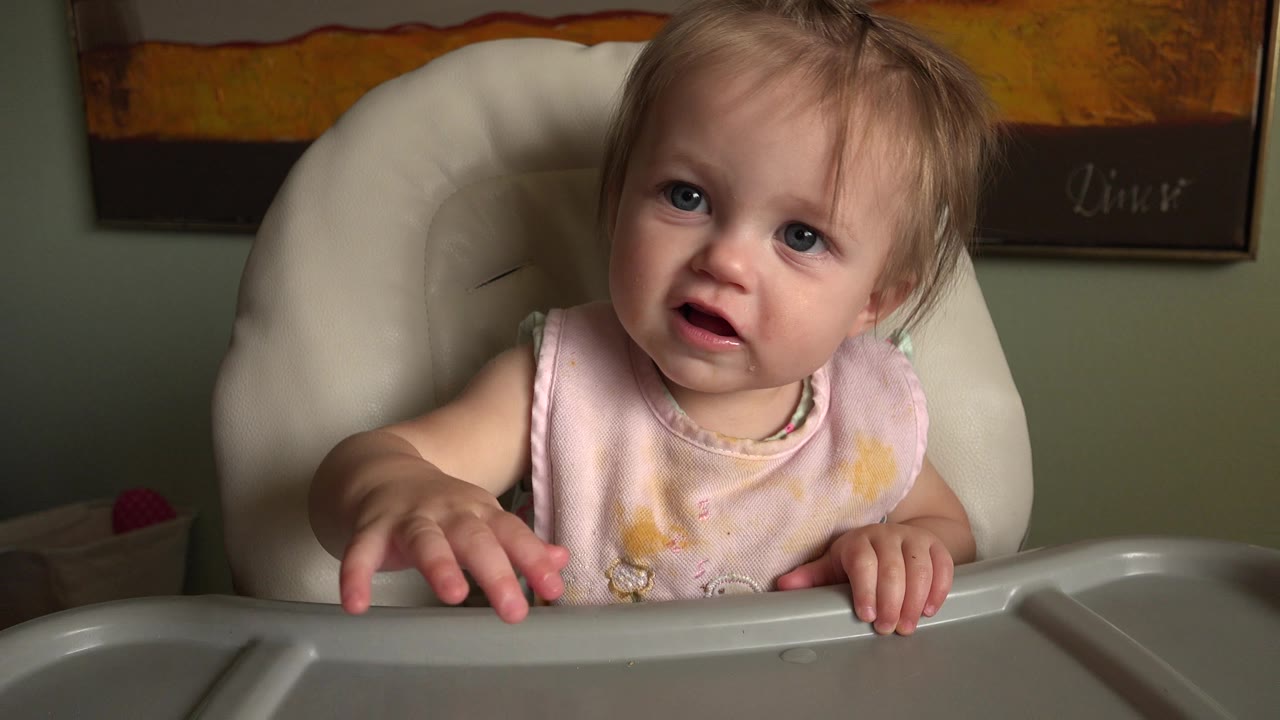 A toddler girl in a pale pink bib sits in a cream high chair at Hidden Lake, Colorado, with one hand on the gray tray and framed artwork on the green wall behind her.