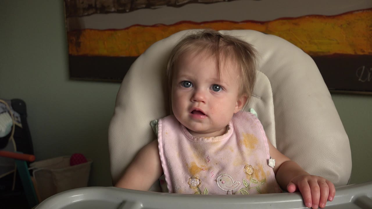 A blonde baby in Hidden Lake, Colorado sits centered in a white highchair wearing a pale pink weaning bib, with a framed wall artwork behind.