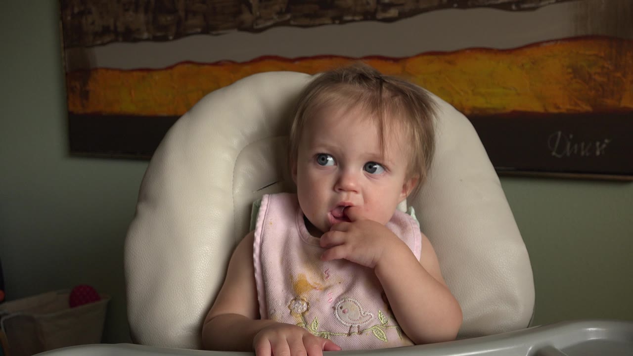 Toddler in a pink bib sits in a white padded highchair.
