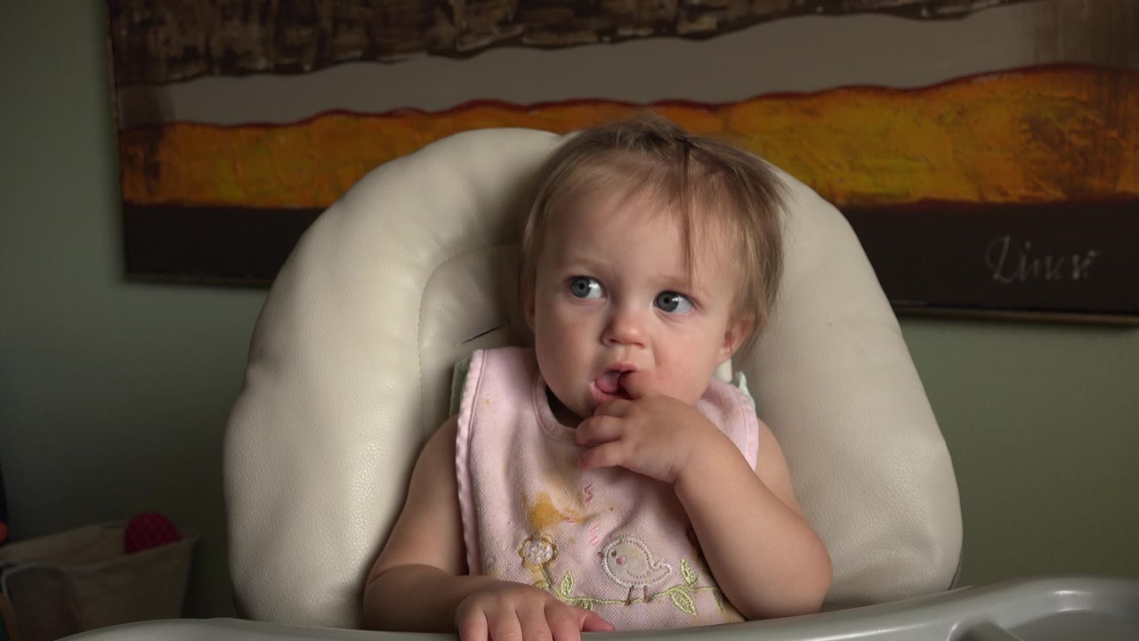 At 0:01, the toddler remains in the white padded highchair in Hidden Lake, Colorado, looking off to the side with one hand near the mouth and the tray visible below.