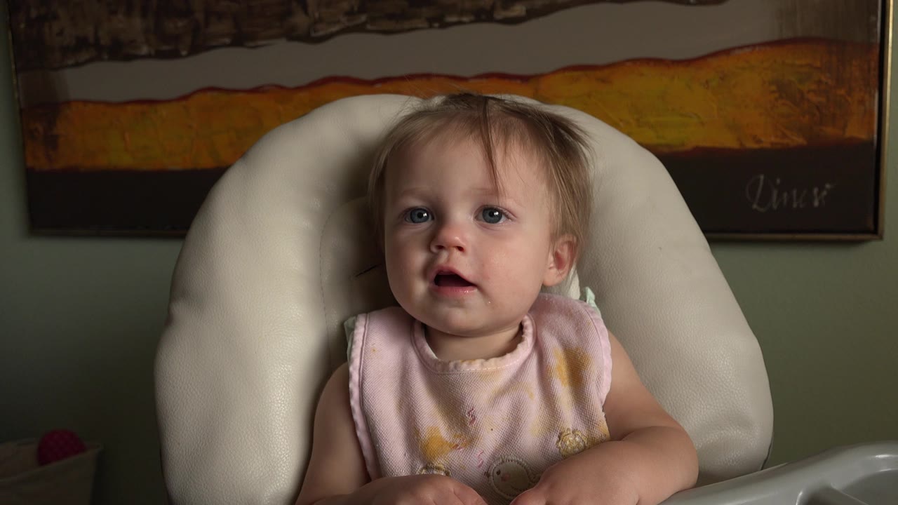 At 0:01, the child remains centered in the white high chair, facing forward with a slightly open mouth and the warm-toned wall art visible behind.