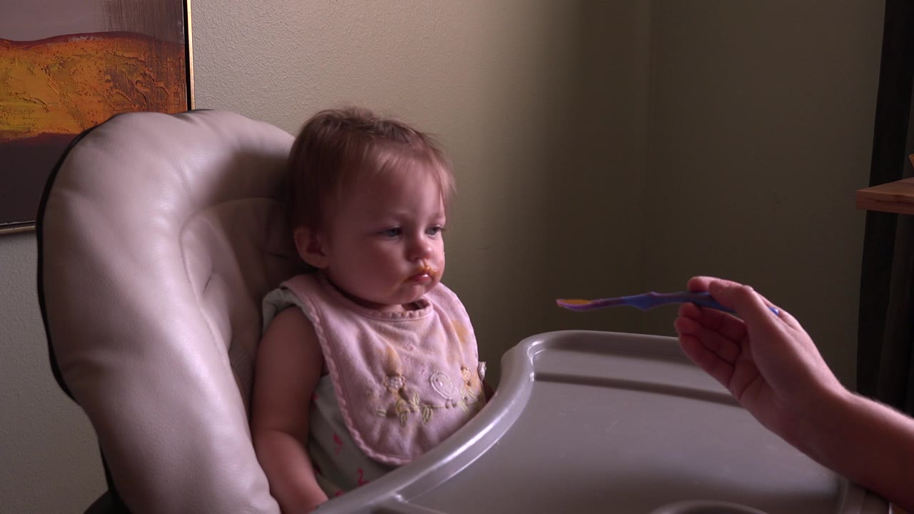 Baby in high chair faces a spoon held by an adult hand.