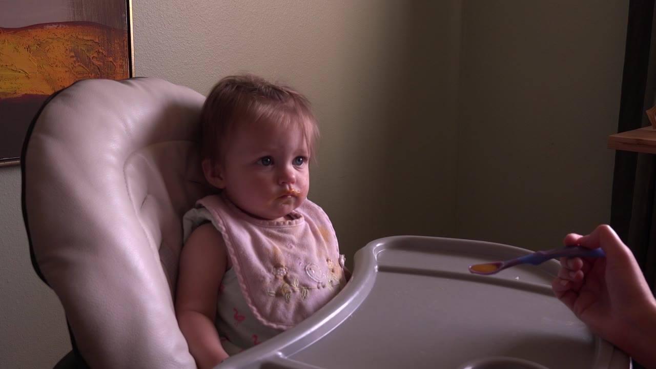 Baby in a high chair looks toward a spoon held at the tray.