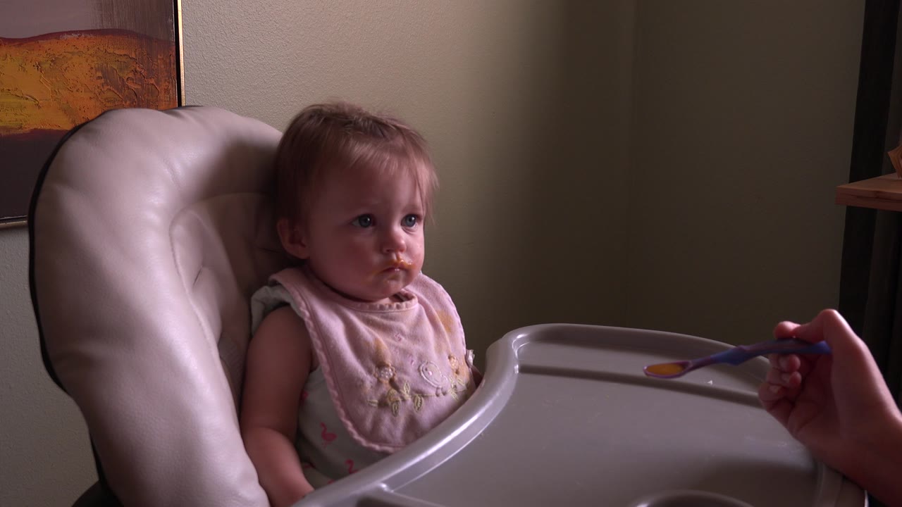 At 0:01, the baby remains seated in the high chair while the spoon is held from the right side of the frame in the Hidden Lake, Colorado home-movie clip.