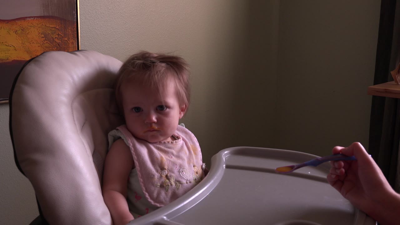 At 0:00, a toddler sits in a pale high chair at Hidden Lake, Colorado, with an adult hand holding a spoon near the tray and a wall hanging partly visible behind.
