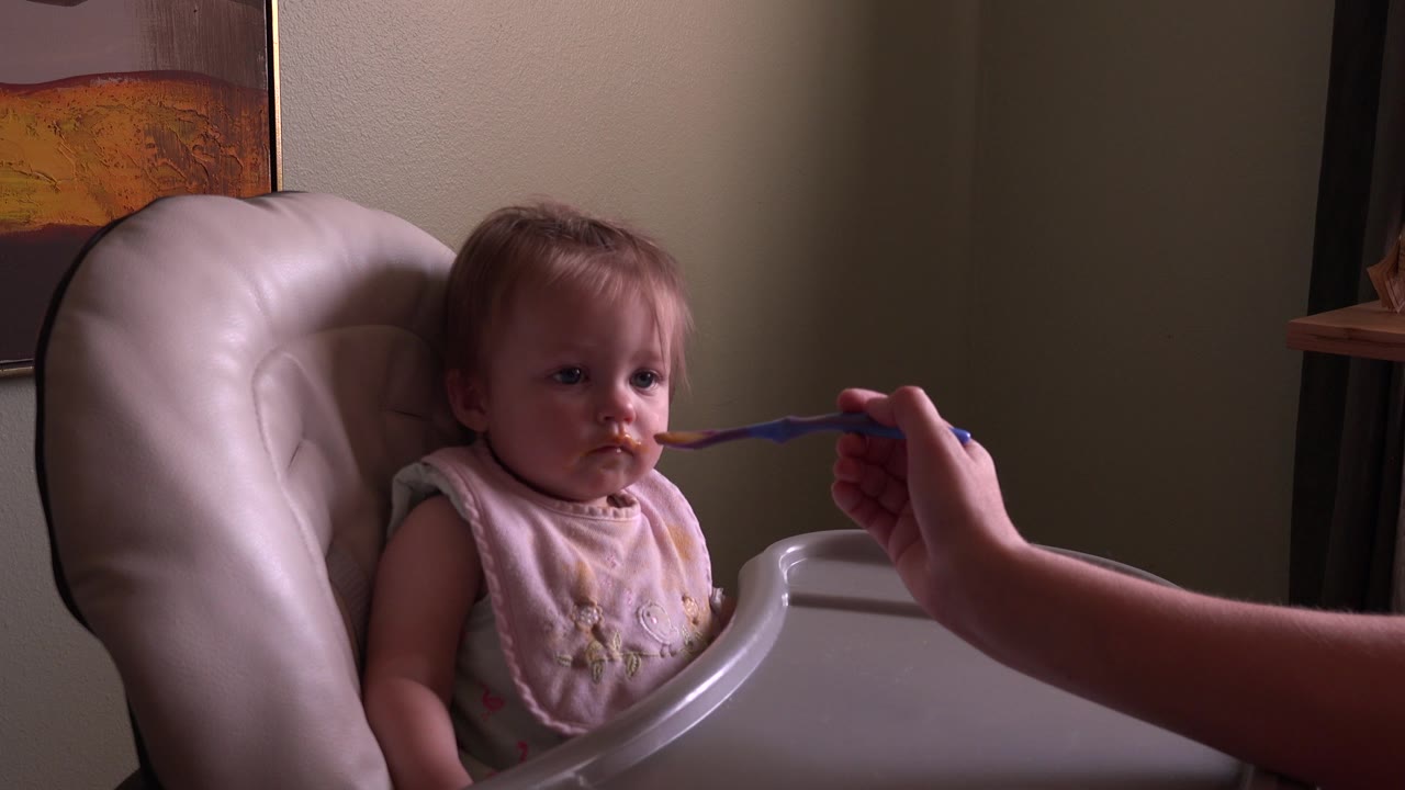 Baby in a white high chair looking at a purple spoon.