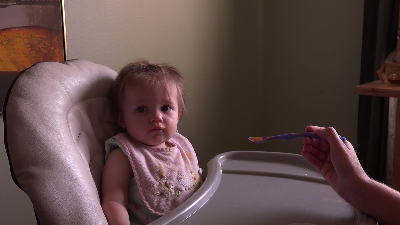 Baby in a high chair faces a spoon held from the right.
