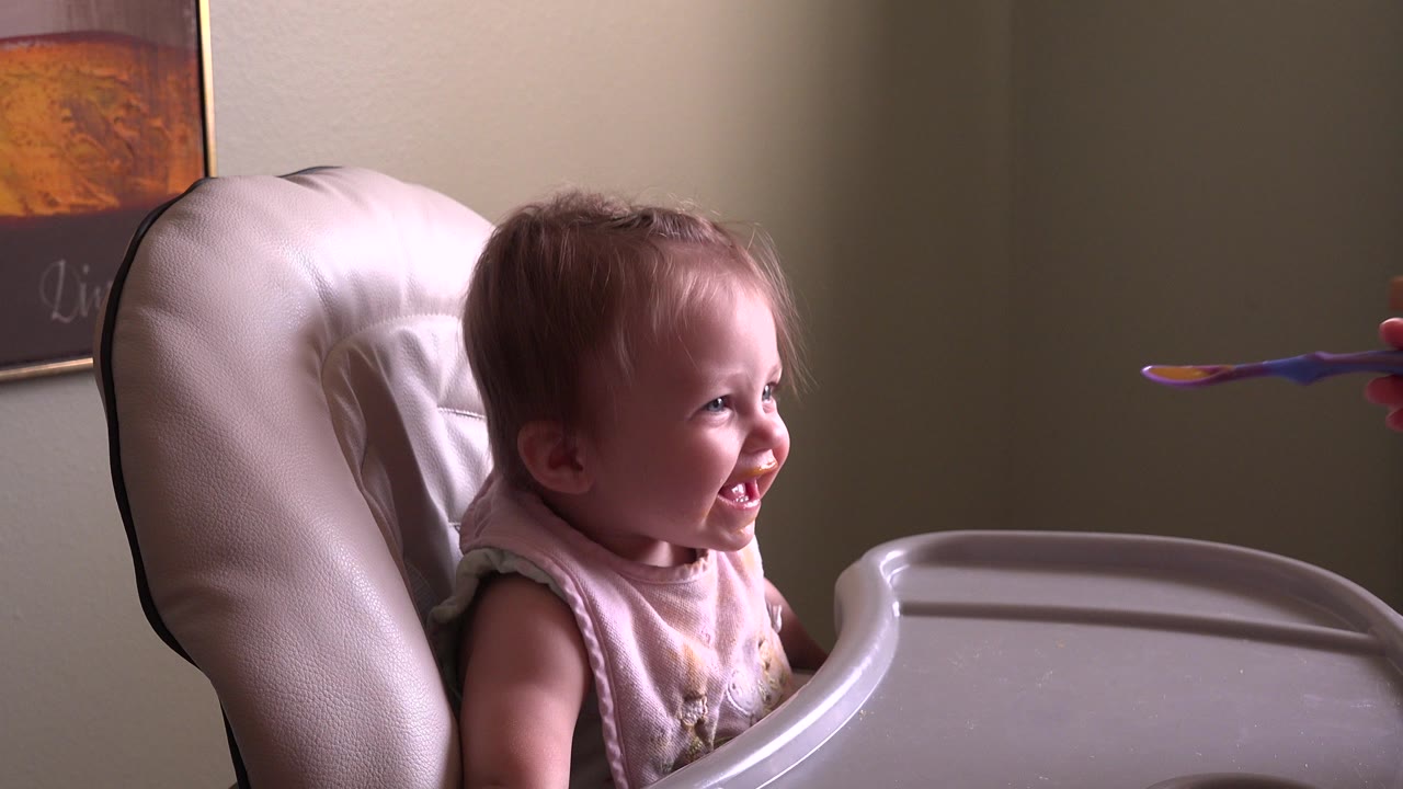 Baby smiling in a high chair as a spoon approaches from the right.