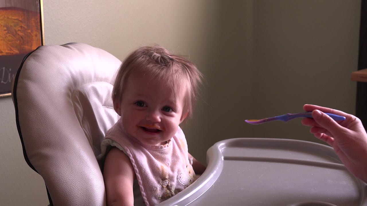 Baby in a high chair facing a spoon held from the right.