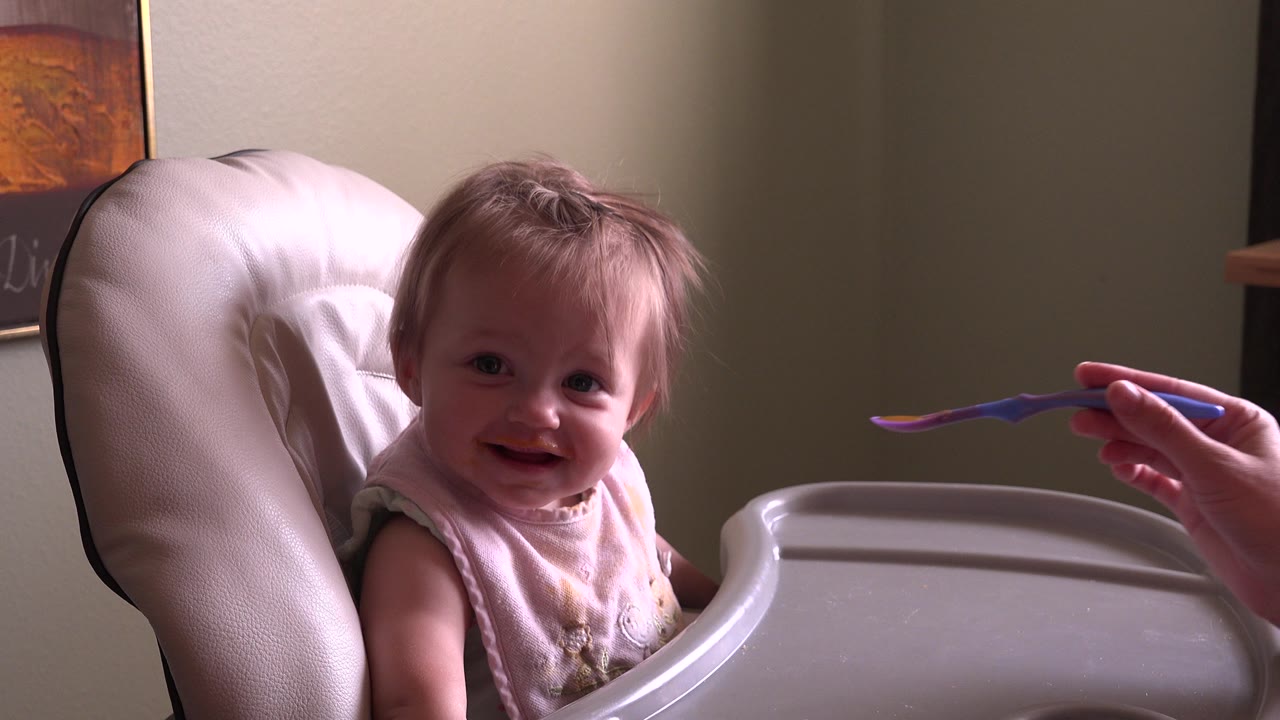 At 0:01 in Hidden Lake, Colorado, the baby looks toward the camera while the adult hand keeps the spoon extended beside the high chair tray.