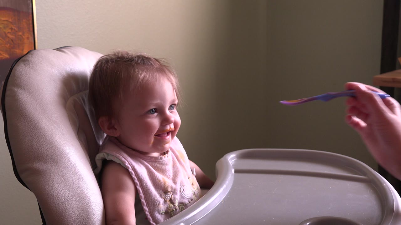 A purple spoon enters from the right toward the smiling baby in the booster chair, with the indoor wall and framed art still visible behind.