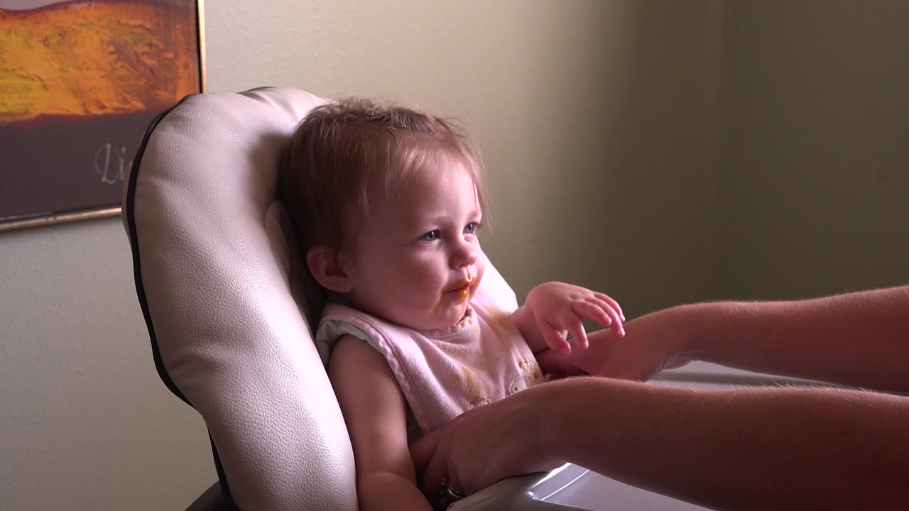 Child in a high chair with food on the mouth as an adult arm reaches in.