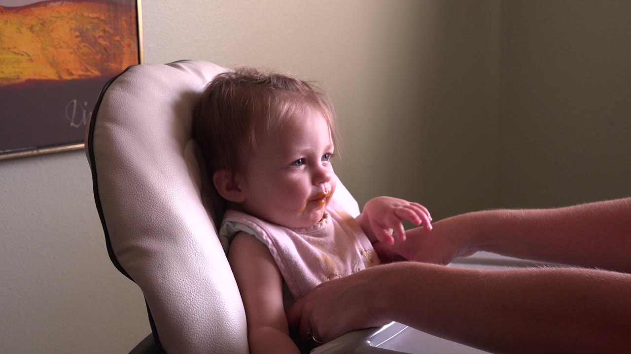 The child remains seated in the high chair at Hidden Lake Colorado, looking toward an adult arm across the tray with a stained bib visible in soft indoor light.