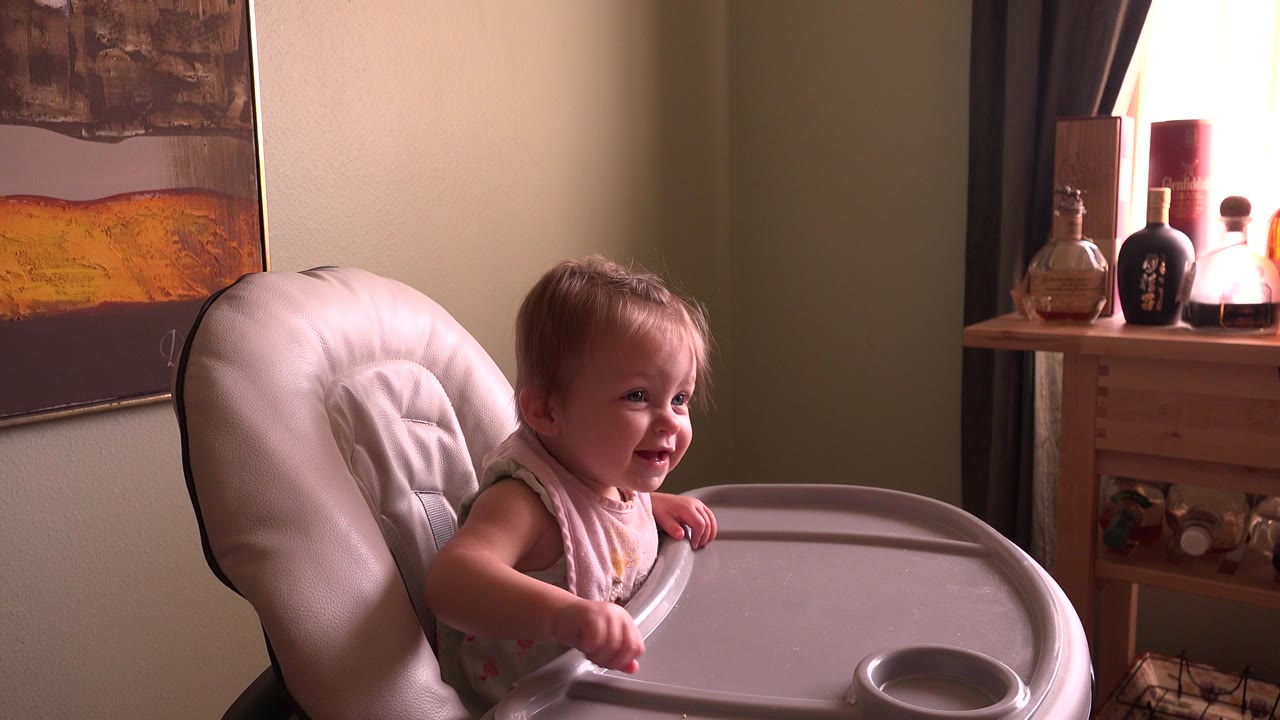 At 0:00, a baby girl in Hidden Lake, Colorado sits upright in a padded high chair, smiling slightly beside a wall painting, curtains, and warm window light.