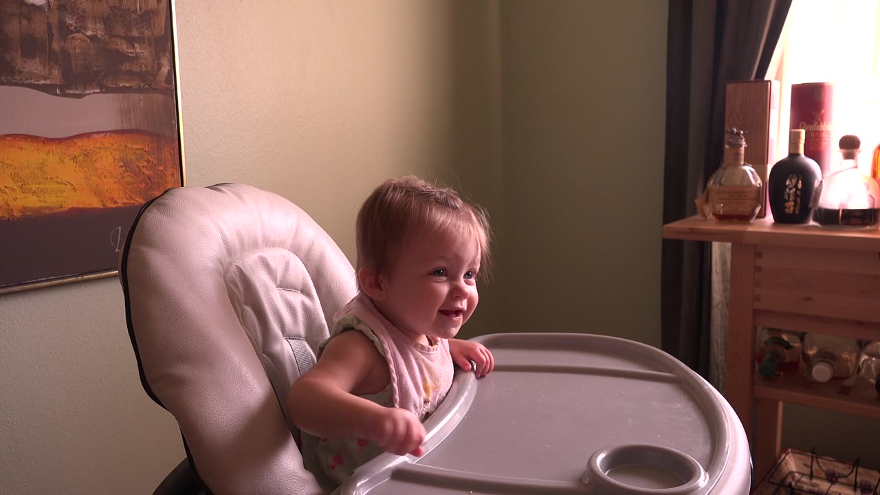 At 0:01, the baby girl in Hidden Lake, Colorado leans near the high chair tray and smiles toward someone off camera, with the side table and window area behind her.