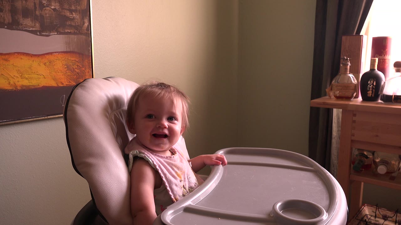 Baby girl smiling in a high chair beside a wall painting and window.
