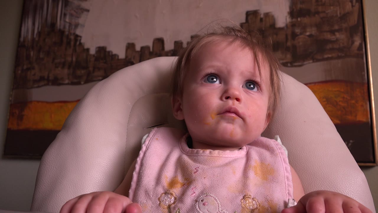 Baby in a padded seat looking upward with food marks on a pink top.