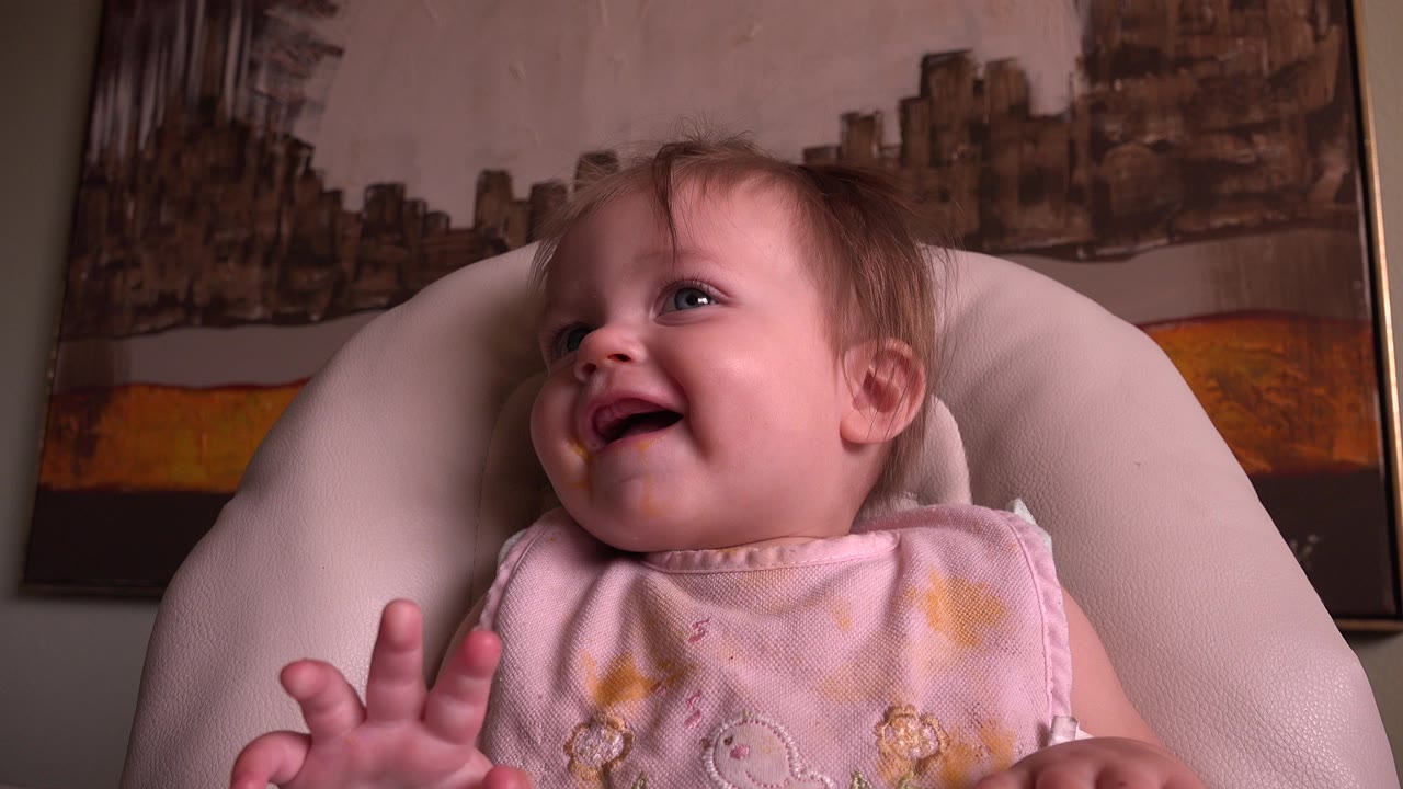 At 0:00 in the Hidden Lake, Colorado clip, a toddler in a pink bib laughs while seated in a pale cushioned chair with framed wall artwork behind.