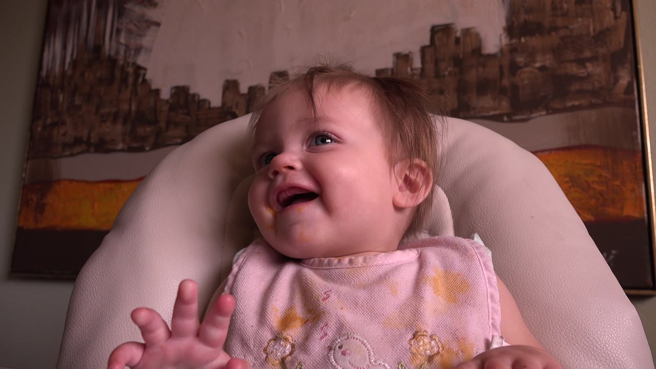 At 0:01, the toddler keeps laughing in the chair, with one small hand raised in the foreground and the wall artwork remaining behind the seat.