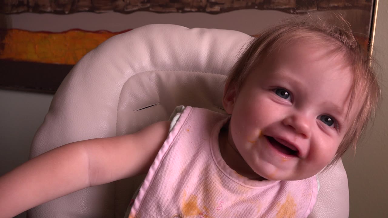 At Hidden Lake, Colorado in 2015, a smiling baby in a white high chair leans toward the camera while wearing a pink bib marked with food stains.