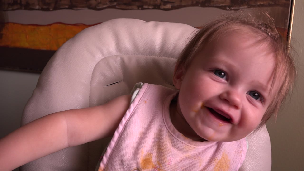 At Hidden Lake, Colorado in 2015, the baby keeps smiling toward the camera from the padded white high chair, with the adult arm still partly in frame.