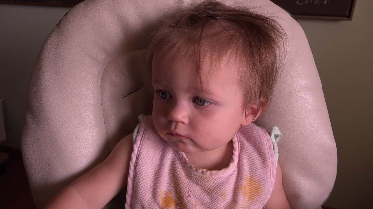 At 0:00, a young girl in Hidden Lake, Colorado sits in a cream padded chair, looking off to the left while wearing a pink sleeveless top.